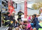 Feuerwehrleute sitzen mit Kindern vor Feuerwehrauto und erklären etwas